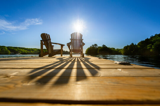 Sunrise on a lake in Muskoka, Canada. Two empty Adirondack chairs sits on a dock on a peaceful lake.