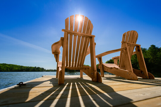 Sunrise on a lake in Muskoka, Canada. Two empty Adirondack chairs sits on a dock on a peaceful lake.