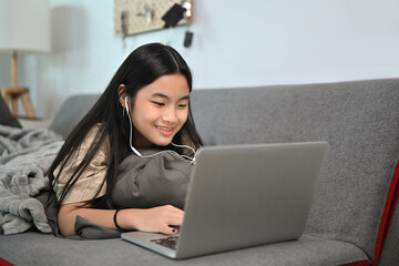 Cheerful young woman lying on sofa and surfing internet with laptop computer.