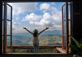 back of Asian woman with freedom arms open standing behind large wooden window with mountain view and cloudy blue sky at Doi Chang, travel attraction in Chiang Rai, Thailand    - Powered by Adobe