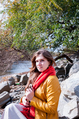 A girl with long hair in a yellow jacket and an orange scarf holds a book sitting on the river bank