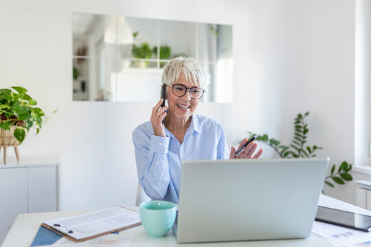 Smiling Mature Beautiful Business Woman With White Hair Working On Laptop In Bright Modern Home Office. Business Woman Talking On Her Mobile Phone While Working From Home