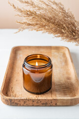 Obraz na płótnie Canvas Cozy burning candle in brown glass jar, winter home interior decor on wooden tray with dry reed bouquet. Eco style.