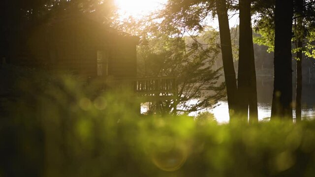 Sun gleams on rustic cabin at top of hill near lake during golden hour (60fps)