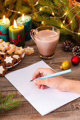 Child writes the letter on a blank clean sheet of paper. Child's hands, the sheet of paper and Christmas decorations on a wooden table. Concept of New year and Christmas.