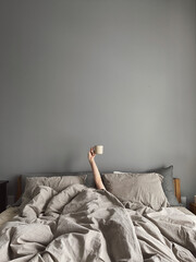 Hand's of young woman with coffee mug in bed with neutral grey bed linen cloth. Minimal happy morning concept