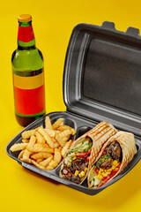 Vegan burrito in takeaway container with fries and bottle of drink on yellow background