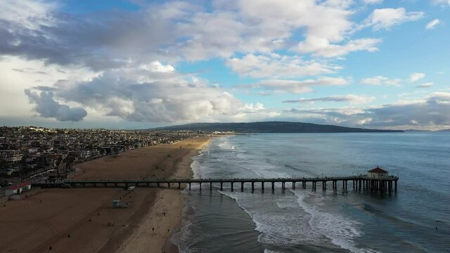 View of a pier with a house on the coast in front of a mountain overlooking the sea, zoom. Drone sunset view of Manhattan Beach, California.  Cloudy skies.