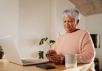 Elderly multi-ethnic female happily making online purchase on laptop, sitting at kitchen counter. 