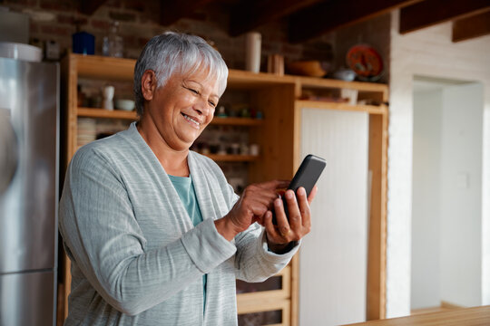 Content multi-cultural elderly female typing a message on smartphone while standing in modern kitchen.