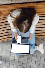 Top view, close-up of smartphone with blank screen in hands of young woman sitting at wooden bench and touching screen. Laptop with blank screen. Mock up space for ad.