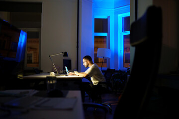 Workaholic man staying in office overnight preparing for business presentation or startup launch. Busy programmer or web content editor working on laptop sit alone at dark workplace. Overtime concept