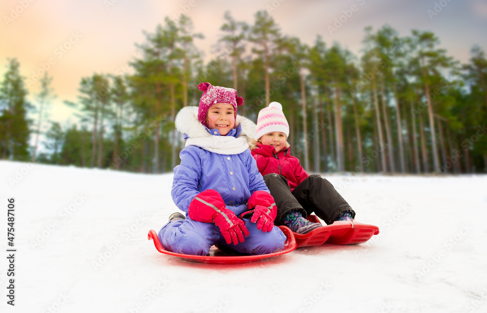 Sticker childhood, sledging and season concept - happy little girls on sleds outdoors in winter over snowy forest or park background