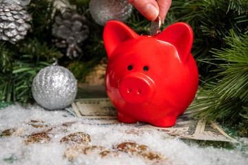 Hand putting a coin into a red piggy bank on gold coins and paper money covered with snow and...