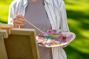 creativity, art and hobby concept - - close up of senior woman with easel and color palette painting outdoors