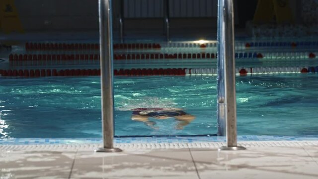 Young woman swimmer in red swimsuit swims to the ladder of a pool and takes off the glasses