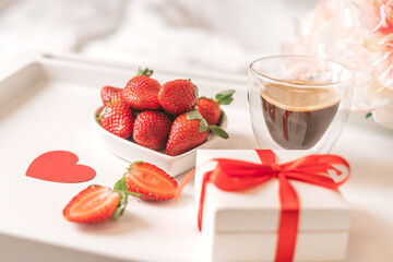Breakfast for Valentine's Day. Heart shaped white plate with fresh strawberries, cup of coffee, gift and flowers with gift in bed. Still life composition.