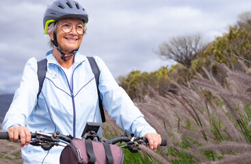 Fototapeta premium Smiling senior woman riding in a country road with her bike, cyclist wearing sport helmet and backpack