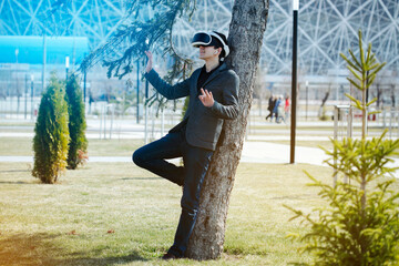 Young  man dressed in virtual reality goggles in a city park near green young spruces and trees, leaning against a tree and manipulating his hands, from behind strongly unwinding background