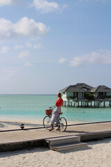 Fototapeta premium A girl in a red blouse and a white skirt stands with a bicycle on a wooden pier leading to villas on stilts above the water in the Maldives.