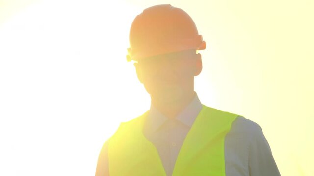 Engineer in uniform at substation. Specialist of maintenance in orange hardhat and yellow vest walks against bright sunlight closeup