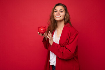 Portrait of positive cheerful fashionable woman in formalwear holding small money box looking like red pig looking at camera isolated on red background with copy space