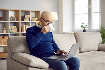 Middle-aged businessman sit on sofa at home work online on laptop drink morning coffee. Pensive senior male worker use computer communicate on internet. Distant remote job, freelance concept.
