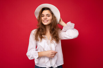 Portrait of young attractive caucasian hipster woman in trendy casual clothes and white hat. Sexy carefree female person posing isolated near red wall in studio. Positive model with natural makeup