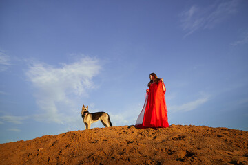 Beautiful young woman in red dress with white wings on the sand and dog german shepherd on sunny day with blue sky. Angel model or dancer posing in photo shoot on dunes