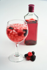 Balloon glass full of ice and raspberries and blackberries gin with a bottle of red gin on a white baackground