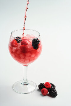 Pouring raspberry and blackberry gin into a full balloon glass with raspberries and blackberries in it on a white background