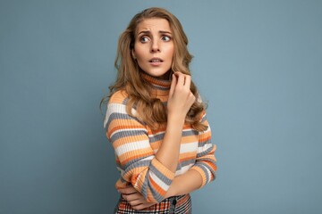 Portrait photo of young pretty beautiful attractive scared asking thinking blonde woman with wavy-hair wearing striped sweater isolated over blue background with empty space for text