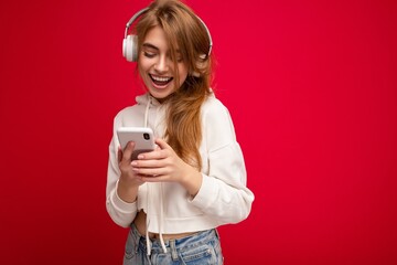 Photo of beautiful happy smiling young blonde woman wearing white hoodie isolated over colourful background wall with copy space for text wearing white wireless bluetooth earphones listening to cool