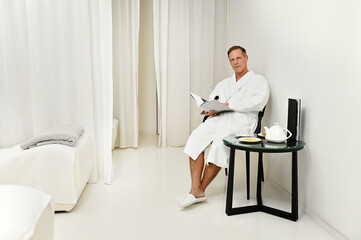 Confident portrait of a handsome mature Caucasian man looking at camera, sitting on an armchair with a magazine in his hands while relaxing in private room of a luxurious wellness spa centre