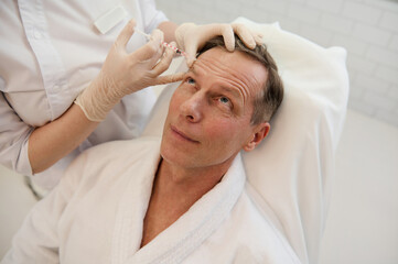 Fototapeta na wymiar Beautician injects a botulinum toxin into a man's forehead to relax the muscles. Mature man receiving facial rejuvenation injection procedure. Men's cosmetology.