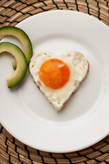 heart-shaped scrambled eggs on a white plate with avocado slices on a jute napkin