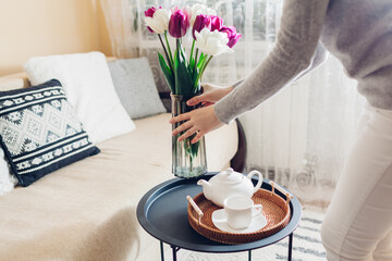 Woman puts vase with tulips flowers on table. Housewife taking care of coziness at home. Interior and spring decor