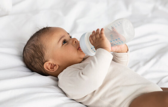 Cute little black baby drinking from bottle lying in bed