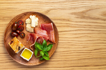 Italian ham, parmesan cheese, focaccia, tomato and basil twig in a wooden plate on a wood background with copy space. Light Italian antipasti. Flat lay. Top view.