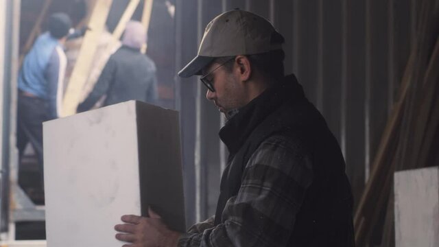 Tilt up view of mature male artisan in casual clothes with glasses and cap touching and inspecting block of polymer near entrance of hangar during work - Powered by Adobe