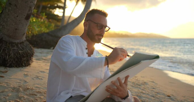 Handsome artist painting wonderful seascape of the evening tropical ocean sitting under palm trees