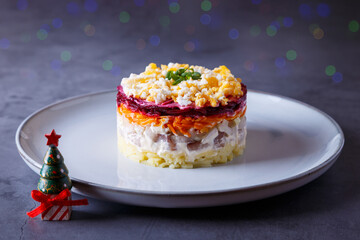 Herring salad under a fur coat. Traditional Russian multilayered salad from herring, beets, potatoes, carrots and eggs. Close-up, grey background, miniature Christmas tree, bokeh.
