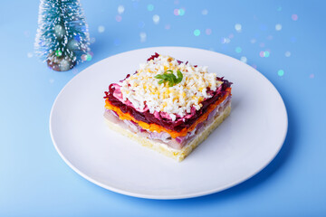 Herring salad under a fur coat. Traditional Russian multilayered salad from herring, beets, potatoes, carrots and eggs. Close-up, blue background, miniature Christmas tree.