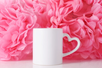 White mug on a pink paper flowers background with heart shaped handle for Valentine's day or Mothers day, template for your design.
