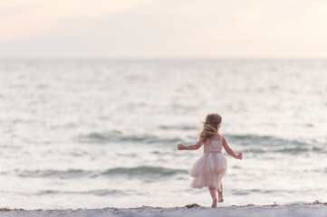 Fototapeta na wymiar A Young Girl Kid Child Dancing Jumping Walking Running in a Pretty Pink Dress Water Sand Sunset Sun Sun light Beach Horizon Fun Get Outside Childhood Unplugged