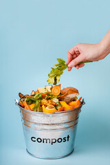 Woman puts kitchen waste in compost-bucket on blue background. Compost-container. Sustainable lifestyle. Vegetable, fruit peels, scraps from food preparation collected in trash-can for recycling