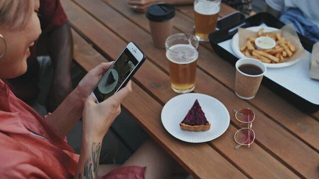 Medium close up of young Asian woman taking photo of delicious piece of cake on smartphone while sitting at outdoor cafe with friends on summer evening