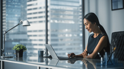 Successful Businesswoman in Stylish Dress Sitting at a Desk in Modern Office, Using Laptop Computer, Next to Window with Big City View. Successful Finance Manager Planning Work Projects.