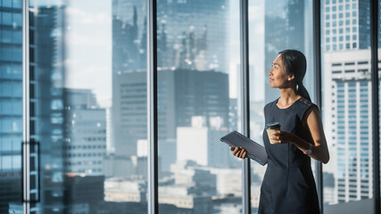 Successful Businesswoman in Stylish Dress Standing in Modern Office, Holding Laptop and Drinking...