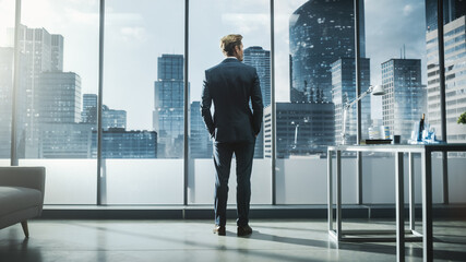 Fototapeta na wymiar Thoughtful Young Businessman in a Perfect Tailored Suit Standing in His Modern Office Looking out of the Window on Big City with Skyscrapers. Successful Finance Manager Planning Project Strategy.